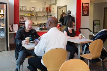 Two males sat in a coffee shop, talking and laughing 