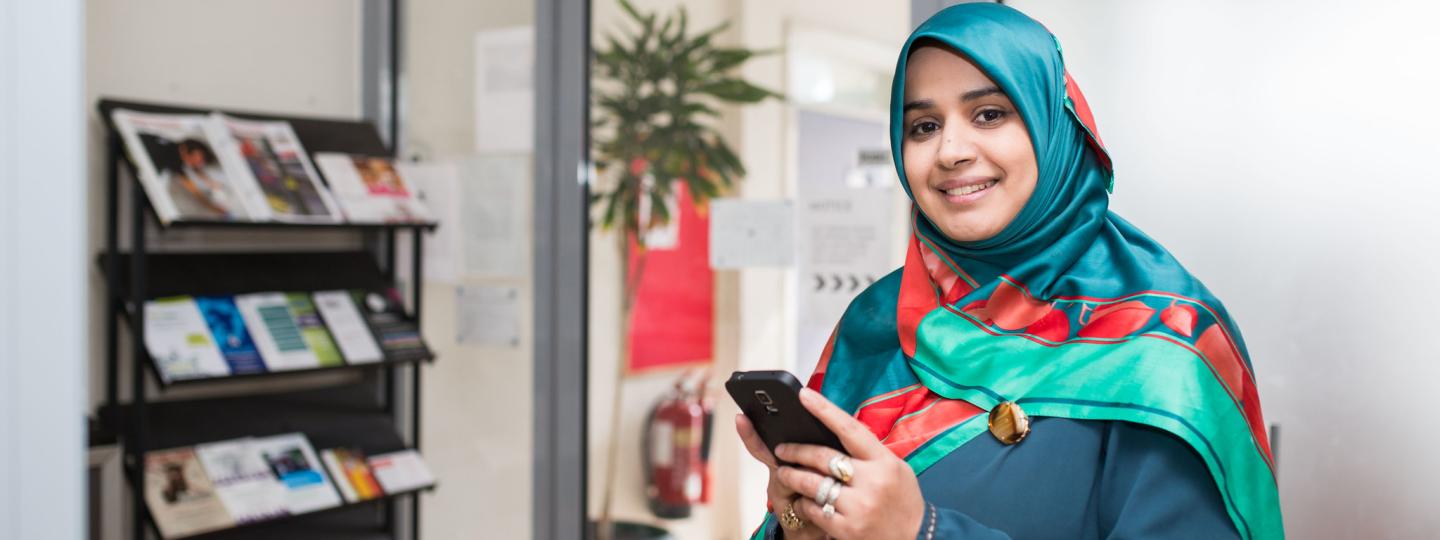 Smiling woman wearing a hijab, holding out a phone.